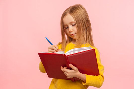Child doing homework. Portrait of clever little girl writing poem in book with serious concentrated expression, development of creative child abilities. indoor studio shot isolated on pink background