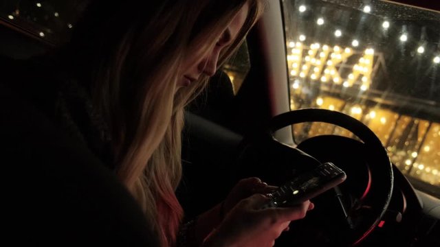 Blonde in a car with a smartphone in her hands. Girl is typing closeup. Night city. Slow motion.