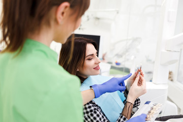 Dentist shows crown samples to his patient. Woman at the dentist's appointment chooses the tone of...