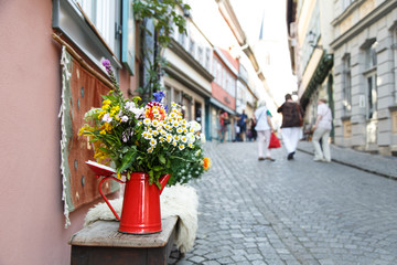 summer bouquet in red antique milk jug on a bench. In the background in the blur a street with historic houses and groups of visitors
