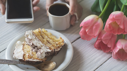 Tiramisu cake, female hands holding smartphone and cup of coffee and pink tulips