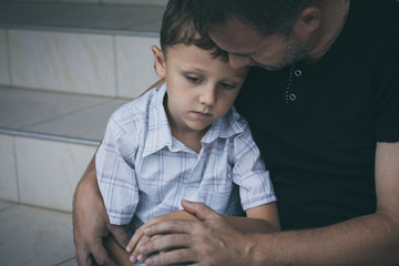 Portrait of young sad little boy and father sitting outdoors at the day time.