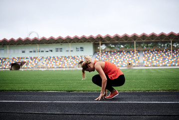 Young blond sportswoman, wearing orange top and black leggings, is squatting down and leaning forward, stretching her back. Warming up before fitness training on stadium with green grass in summer