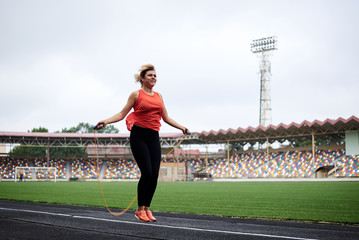 Fototapeta premium Full-length picture of sportswoman, jumping on jump rope on stadium. Young blond woman, wearing orange top and black leggings, doing fitness training in the morning in summer.