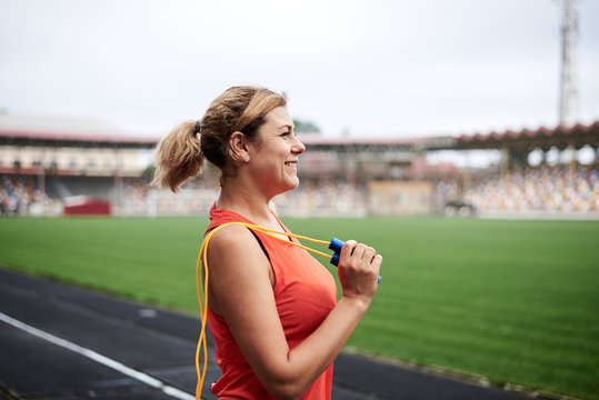 Young blond woman, wearing orange top, holding yellow jump rope on her shoulder. Three-quarter profile picture of sportswoman on stadium. Fitness training in the morning in summer. Exercise outdoors