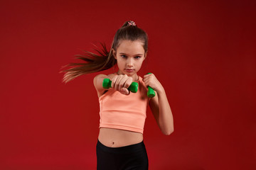 Fototapeta na wymiar Refuse to Lose. A cute kid, girl is engaged in sport, looking at camera while exercising with dumbbells. Isolated on red background. Fitness, training, active lifestyle concept. Horizontal shot