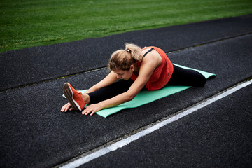 Young blond woman, wearing orange shirt and black legging sitting on light green yoga mat, in semi-twine, stretching her back, legs, and body. Yoga fitness exercise outside on stadium with green grass