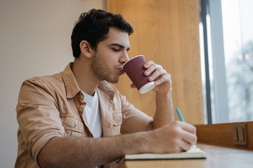 Indian business man drinking coffee in cafe, taking notes, planning start up. Freelancer working...