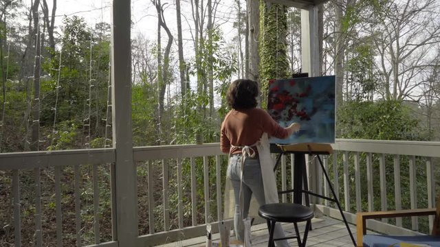 Young professional female painter works on abstract multicolored painting with brush on canvas outside at sunset, camera dolly right