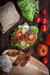 closeup of smoked meat sandwich with green salad, fresh tomatoes, pickled cucumbers and bread, on dark wooden table background