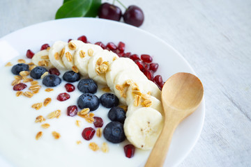 Natural white yogurt on white plate with banana, blueberry,promegranet seed,glanola and cherry on white wooden table.Close up.