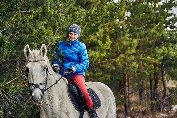 Young woman rider in a blue blazer and sporting a cap for a walk on a white horse on a cloudy winter day.