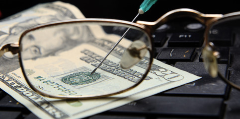 On the computer keyboard is a twenty-dollar bill. A medical syringe is inserted into the surface of...