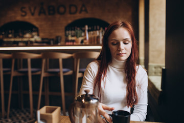 Close up portrait of young attractive ginger girl with freckles sitting i a cafe drinking coffe alone, looking in the window. Emotional concept, freckled people concept