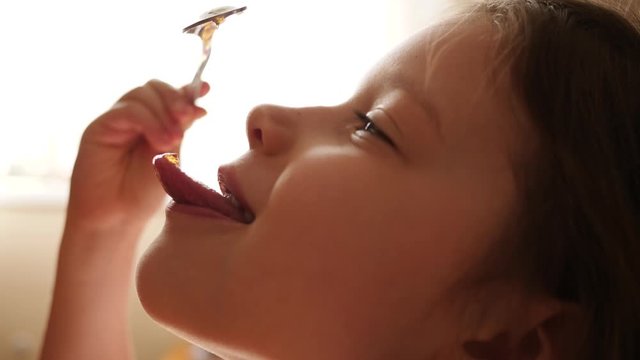 Little child girl eat honey as it drops on her tongue licking it out from the spoon