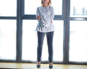 Obraz premium Portrait of a business woman in a white elegant blouse standing against the background of large windows on the floor. Business, elegant business woman. Interior. Telework.