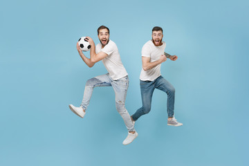 Fototapeta na wymiar Excited two young men guys friends in white t-shirt isolated on pastel blue background in studio. Sport leisure lifestyle concept. Cheer up support favorite team with soccer ball jumping like running.