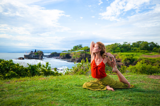 Outdoor yoga practice. Young woman practicing Eka Pada Rajakapotasana, One Legged King Pigeon Pose. hest opener improving breathing. Tanah Lot temple, Bali, Indonesia