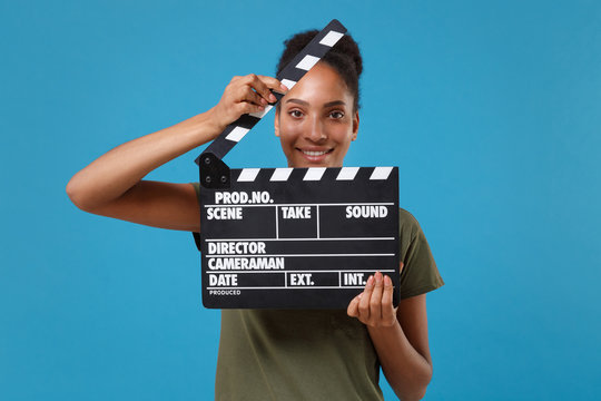 Smiling young african american woman girl in casual t-shirt posing isolated on bright blue background. People lifestyle concept. Mock up copy space. Holding classic black film making clapperboard.