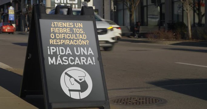 A sign outside of a store in a city's business district informs customers and pedestrians to ask for a face mask to help the spread of Coronavirus COVID-19. Spanish version.  	