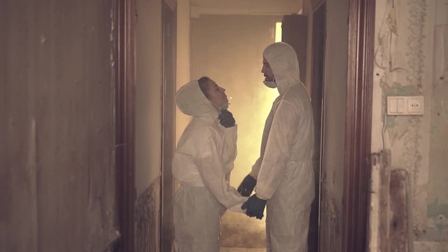 Slow motion young couple wearing  insulation bio hazard suit and gear remove their face masks and kiss in the middle of an abandoned house