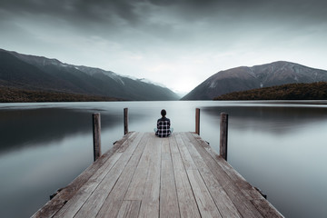 A woman looking at the horizon on the rototiti lake bridge in the Tasman region, in Nelson Lakes national park, facing the lake and the mountains during bad weather - obrazy, fototapety, plakaty
