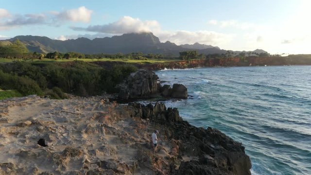 Aerial video of a man hiking down along a rugged sea cliff by the ocean with waves breaking, mountain range in the background and ironwood pine, Makawehi Lithified bluff, Kauai