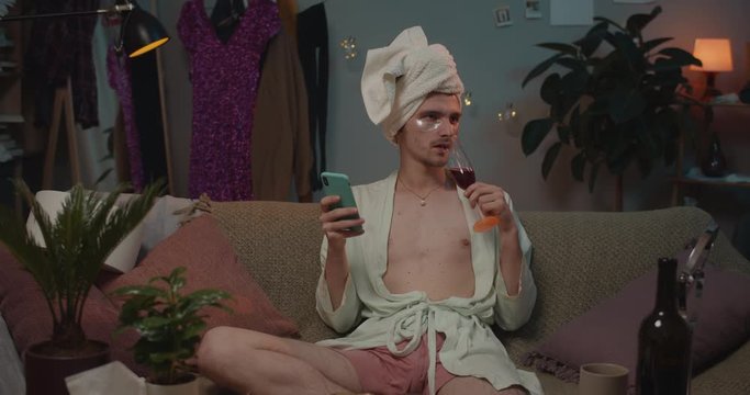 Front view of transgender in rob and towel on head drinking wine and sitting on sofa with smartphone. . Man transgender making jealous face expression while using phone. Home background.