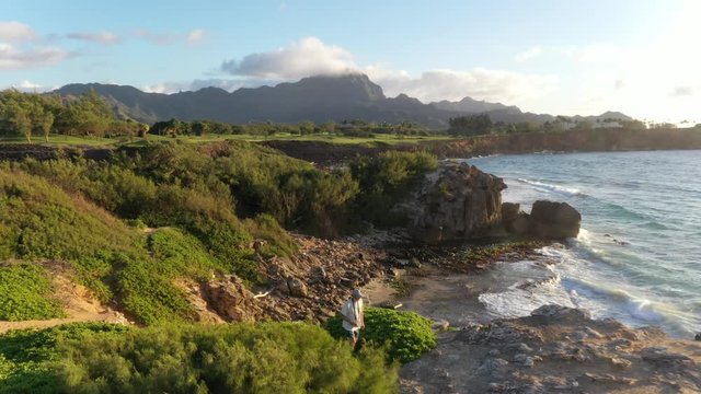 Aerial video of a man hiking right along a rugged sea cliff by the ocean with waves breaking, mountain range in the background and ironwood pine, Makawehi Lithified bluff, Kauai