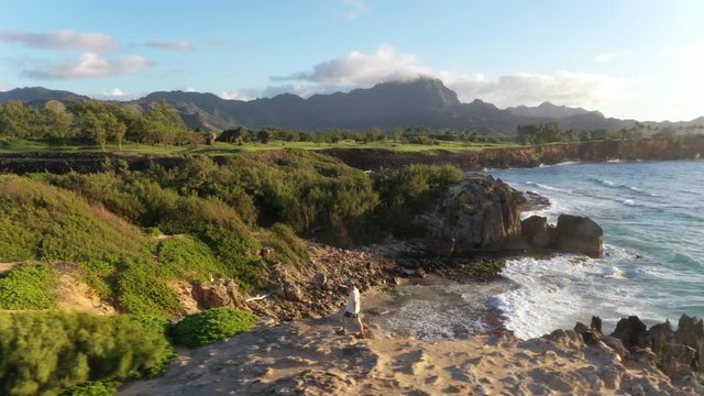 Aerial video of a man hiking down and left at sunrise along a rugged sea cliff by the ocean with waves breaking, mountain range in the background and ironwood pine, Makawehi Lithified bluff, Kauai