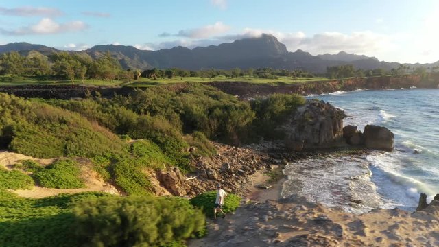 Aerial view of a man running left at sunrise along a rugged sea cliff by the ocean with waves breaking, mountain range in the background and ironwood pine, Makawehi Lithified bluff, Kauai
