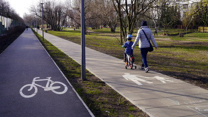 Dziecko na chodniku na rowerze. Obok ścieżka rowerowa. Rowerem po ścieżce rowerowej, po chodniku? Nauka jazdy na rowerze dziecięcym. Znaki informacyjne "droga dla pieszych", "ścieżka rowerowa". 