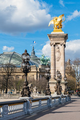 Fototapeta premium Golden statue and lanterns of Pont Alexandre III bridge with Grand Palais and French flag waving on top of the building - Paris, France