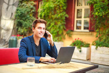 Handsome young business man with mobile phone outdoors at table in summer cafe courtyard working at laptop