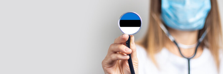 female doctor in a medical mask holds a stethoscope on a light background. Added flag of Estonia....