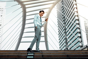 Happy Young Businessman Using Mobile Phone in the City. Lifestyle of Modern People. Low Angle View. Full Length. Looking at Camera and Smiling. Urban City as background