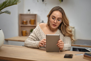 Girl blogger in glasses, beige sweater. Holding tablet, sitting in kitchen at table with notebooks, smartphone, plastic card and palm on it. Close-up