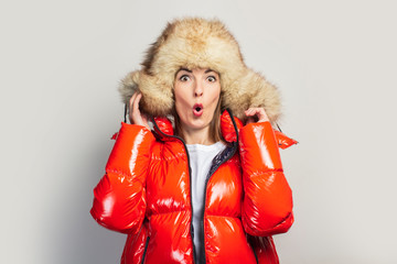 surprised young girl in a red jacket and a fur hat, looking at the camera in shock on a light...
