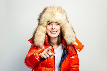 happy young girl in a red jacket and a fur hat, looks at the camera and points a finger in front of...