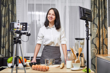 Young woman vlogger recording video for food channel. Female culinary specialist vlogging with her mobile phone mounted on a tripod and ? light stand in kitchen.