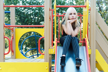Obraz na płótnie Canvas Sad blonde girl sits on slide and thinking about her boyfriend. Unhappy love and dependence concept.
