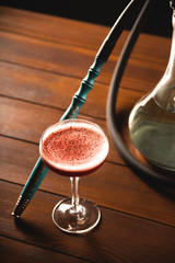 A glass of raspberry cocktail next to a hookah on a wooden table in a dark bar. Alcohol and smoking concept.