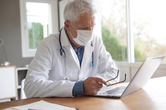 Doctor in office working on laptop, wearing protection mask