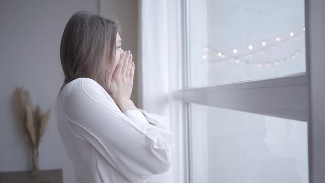 Close-up of sleepy young Caucasian woman yawning and looking out the window in the morning. Side view of pretty brunette girl awakening at home. Lifestyle, leisure, joy.