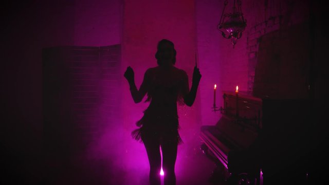 Black mysterious female silhouette with long cigarette mouthpiece in hand short sexy dress old fashioned 20s style. Girl dances incendiary dance. Backdrop room sparkles pink smoke piano burning candle