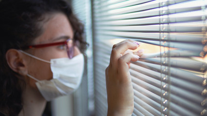 Infected corona virus young beautiful woman or hospital nurse using protective medical face mask look over window blinds