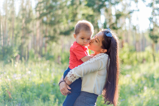 A happy young mother plays and has fun with her young son in nature, in the Park. The mother kisses the baby on the cheek. The concept of a happy family, motherhood. A mother's walk with a small child