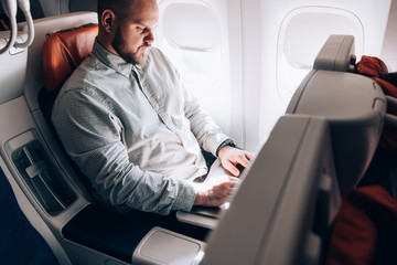 Side view of young Caucasian man using laptop computer near porthole in aircraft connected to wifi on board for chatting online during long trip to other country, male passenger using wireless