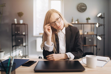 Confident mature lady with blond hair in formal clothing and eyeglasses sitting at table and looking at camera. Business lady posing at modern bright office