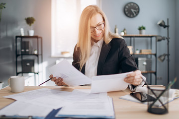 Portrait of concentrated woman in eyeglasses sitting at table in modern office with documents and doing paper work. Mature lady with blond hair in formal clothing skillfully doing her job.